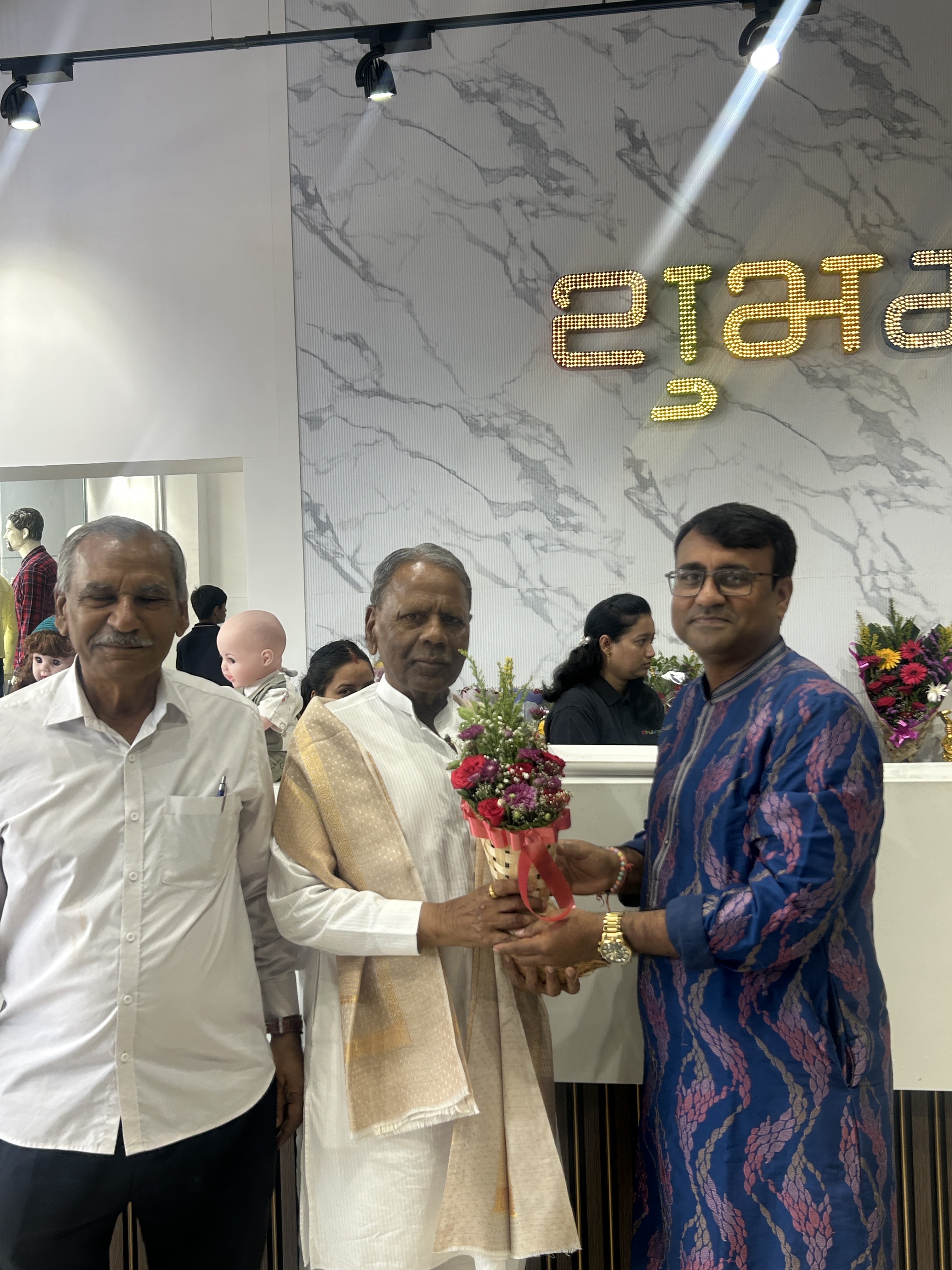Inauguration ceremony at Shubham Fashion store. The owner presenting a bouquet during the ribbon-cutting event, with the शुभम् wall art visible in the background